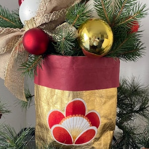 Könnte beinhalten: Ein goldfarbener dekorativer Behälter mit einem rot-weißen Blumendesign. Der Behälter ist mit immergrünen Zweigen, roten und weißen Kugeln und einer goldfarbenen Kugel gefüllt. Ein braunes Band mit goldenem Besatz ist ebenfalls enthalten.