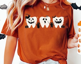 May include: A woman wearing a brown t-shirt with a Halloween-themed design featuring four teeth with spooky faces and black cats. The text "COLOR: AUTUMN" is visible in the top right corner of the image.