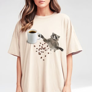May include: Beige t-shirt featuring a raccoon reaching for a white coffee mug filled with dark coffee. Scattered coffee beans are falling from the mug. The raccoon is in mid-air, with a happy expression.