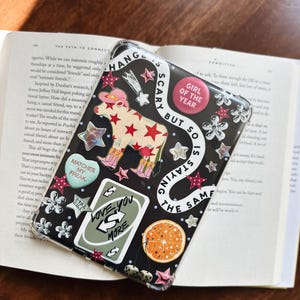Pode incluir: Uma capa protetora transparente para tablet decorada com vários adesivos, incluindo uma vaca com um chapéu de cowboy rosa, um cartão verde com o texto LOVE YOU MORE, uma fatia de laranja, dados e o texto CHANGE IS SCARY BUT SO IS STAYING THE SAME.