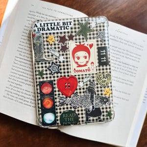May include: A clear protective case for an e-reader or tablet featuring a black and white gingham pattern decorated with various stickers including a cowboy boot, a red tomato character, a traffic light, a heart button, and a fish illustration.
