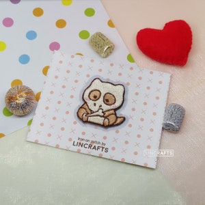 Puede incluir: Un parche termoadhesivo con un personaje de dibujos animados con cuerpo beige claro, ojos marrones y contorno marrón. El parche está sobre una tarjeta blanca con lunares rosas y el texto "Iron-on patch by LINCFRAFTS". Un corazón rojo y carretes dorados y plateados también son visibles.