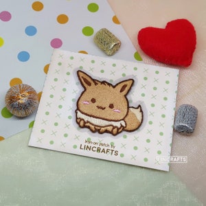 以下が含まれることがあります： 茶色とベージュの刺繍が施されたアイロン接着パッチで、漫画のイーブイポケモンが描かれています。パッチは、緑と白のドット模様の白いカードに貼られており、"Iron-on patch by LINCRAFTS"というテキストが書かれています。