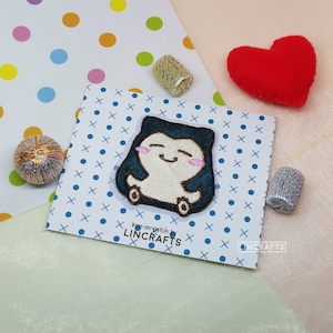 May include: Iron-on patch featuring a cartoon character with a blue hooded cloak, white body, and pink cheeks. The character is smiling and has a happy expression. The patch is on a white background with blue and white polka dots.