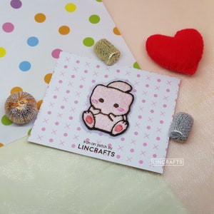 May include: Pink iron-on patch of a cartoon bunny with a pink nose and pink paws. The patch is on a white background with pink and white polka dots. The text "iron-on patch by LINCRAFTS" is printed below the patch.