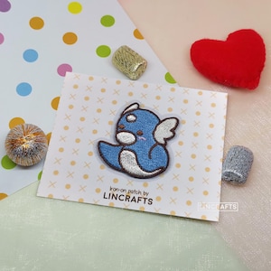 May include: Blue iron-on patch of a cartoon character with wings. The character has a white belly and brown eyes. The patch is on a white card with a yellow polka dot pattern. The text "Iron-on patch by LINCRAFTS" is printed on the card.
