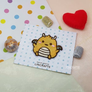 May include: Iron-on patch featuring a cute yellow dragon with a white stripe down its back and pink cheeks. The patch is on a white background with a blue and white cross pattern. The text "iron-on patch by LINCRAFTS" is printed below the patch.
