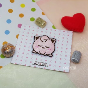 May include: Pink iron-on patch featuring a cartoon character with closed eyes and a curled tail. The patch is on a white background with pink and white polka dots and a white 'X' pattern. The text 'Iron-on patch by LINCRAFTS' is printed below the patch.