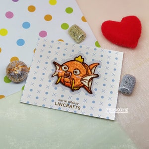 May include: Iron-on patch featuring a cartoon orange fish with a surprised expression. The patch is on a white card with a blue and white pattern. The text "Iron-on patch by LINCRAFTS" is printed on the card.