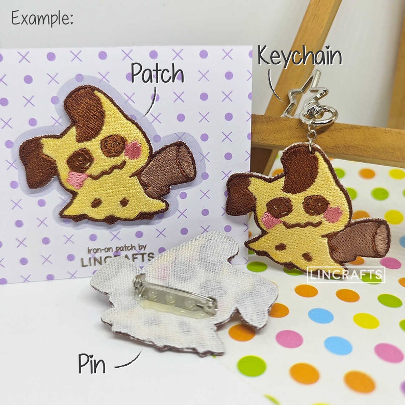 May include: A yellow and brown embroidered patch of a cartoon character with a sad expression. The patch is available as an iron-on patch, a pin, and a keychain. The text "Iron-on patch by LINCRAFTS" is printed on the card. The text "Keychain" and "Pin" are handwritten on the card.