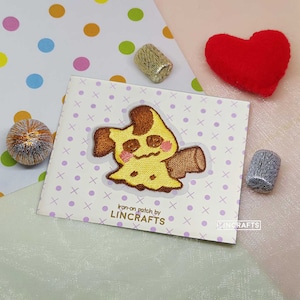 Puede incluir: Parche para planchar con un personaje de dibujos animados amarillo y marrón con una expresión triste. El parche está en una tarjeta blanca con lunares morados y el texto "Iron-on patch by LINCRAFTS".