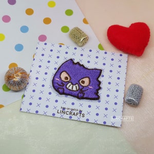May include: Purple iron-on patch featuring a cartoon character with a toothy grin. The patch is on a white card with a blue and white polka dot pattern. The text "Iron-on patch by LINCRAFTS" is printed on the card.