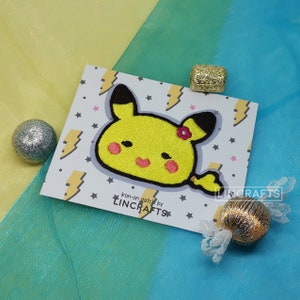 May include: Iron-on patch featuring a yellow cartoon character with a pink flower on its head. The character has a smiling face and a tail. The patch is on a white card with a lightning bolt and star pattern. The text "Iron-on patch by LINCRAFTS" is printed on the card.