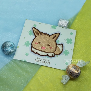 May include: A brown and white embroidered iron-on patch of a cartoon Eevee with a pink tongue sticking out. The patch is on a white card with green shamrock designs and the text "iron-on patch by LINCRAFTS" and "LINCRAFTS".