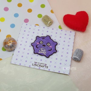 May include: Purple iron-on patch featuring a cartoon character with a skull and crossbones. The character has a cute, friendly expression with large eyes and a smile. The patch is labelled "Iron-on patch by LINCRAFTS".