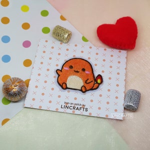 May include: An orange embroidered patch featuring a cartoon character with a flame on its tail. The patch is on a white card with a pink dotted pattern. The text "Iron-on patch by LINCRAFTS" is printed on the card.
