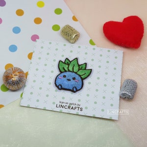 Peut inclure: Patch thermocollant représentant un personnage de dessin animé bleu et vert avec une expression joyeuse. Le patch est étiqueté "Iron-on patch by LINCRAFTS".