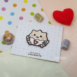 Puede incluir: Parche para planchar con un personaje de dibujos animados en un huevo, con una cáscara rosa y azul. El personaje tiene un contorno marrón y una expresión linda. El parche está en una tarjeta blanca con el texto "Iron-on patch by LINCRAFTS" y "LINCRAFTS" impreso en ella.