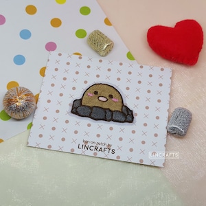 May include: Iron-on patch featuring a cartoon mole with brown fur, pink cheeks, and a small nose, peeking out of a gray dirt mound. The patch is labeled "Iron-on patch by LINCRAFTS".