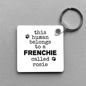 May include: A square white keychain with the text "this human belongs to a FRENCHIE called rosie" in black. The keychain has a silver keyring attached. Paw prints are used as design elements.