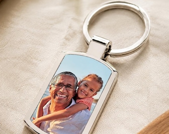 May include: A rectangular silver keychain with a photo of a smiling man and a young girl. The keychain has a rounded top and a split ring for attaching keys. The photo shows the man wearing glasses and the girl hugging him.