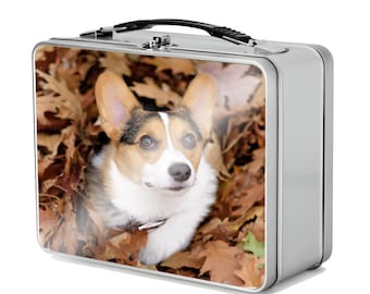 May include: A silver metal lunchbox with a black handle. The lunchbox features a colourful image of a corgi dog lying in a pile of autumn leaves.