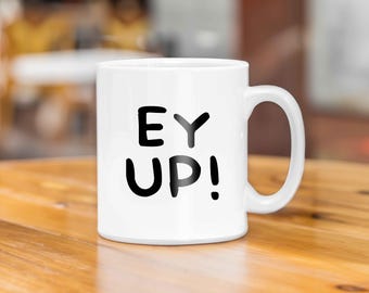May include: White ceramic mug with black text that reads "EY UP!" The mug is sitting on a wooden surface.