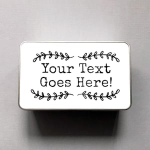 May include: A rectangular, silver-edged tin with a white top. The top features the text "Your Text Goes Here!" in a bold, black font, framed by decorative black leaf sprigs.