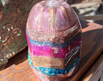 May include: A multi-layered, clear glass orb with a crystal point on top. The orb is filled with layers of glitter, crystals, and other materials in shades of pink, gold, blue, and brown.