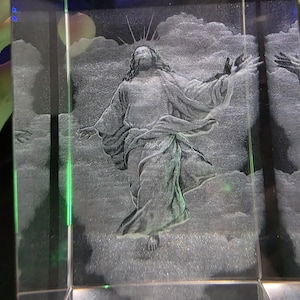 May include: A clear rectangular glass block featuring a three-dimensional laser-etched engraving of a figure with long hair and flowing robes, arms outstretched amidst clouds, with a radiant light effect emanating from behind the head.