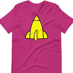 Puede incluir: Camiseta magenta con un diseño de cohete amarillo con contornos negros. El cohete tiene una parte superior puntiaguda y dos aletas laterales. La camiseta está hecha de un material suave y tiene mangas cortas.