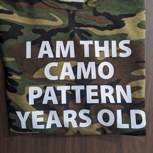 May include: A camouflage green t-shirt with white text that reads "I am this camo pattern years old".