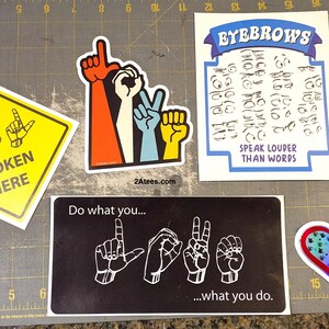 May include: A selection of stickers with diverse designs. One is yellow with "Spoken Here" and hand symbols. Another shows "LOVE" in sign language. Other stickers include a heart and a design with the words "Speak Louder Than Words".
