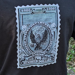 May include: Black t-shirt with a graphic of a vintage-style postage stamp featuring the United States Department of Justice seal and the text "$200 TWO HUNDRED DOLLARS".