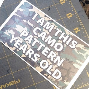 May include: A stack of 12 stickers with a camouflage pattern and the text "I am this camo pattern years old" printed on them. The stickers are on a green cutting mat.