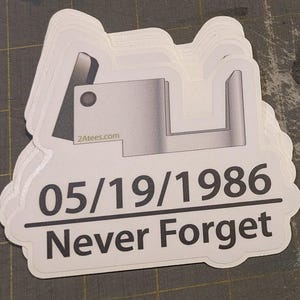 May include: Two white stickers with black text and a gray graphic. The stickers read "05/19/1986 Never Forget." One sticker features a graphic of a camera, and the other has a graphic of a camera component.