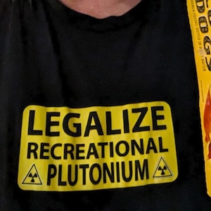 May include: A person wearing a black t-shirt with the text "Legalize Recreational Plutonium" printed on it. The person is holding a box of Foster Farms Corn Dogs. The box is yellow with black text and a picture of a corn dog. The text on the box reads "Corn Dogs, Chicken Franks Dipped in Honey Batter, 7g Protein per Serving, No Hormones".