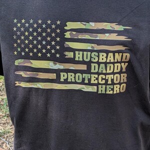 May include: Black t-shirt with a camouflage American flag design and the words "HUSBAND DADDY PROTECTOR HERO". The shirt is displayed on a mannequin in an outdoor setting, with the website 2Atees.com printed on the bottom.