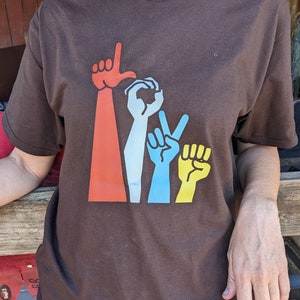 May include: A brown t-shirt with a graphic of three hands forming the letters "I Love You" in sign language. The hands are red, blue, and yellow.
