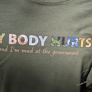 May include: Green t-shirt with a camouflage pattern. The text "MY BODY HURTS and I'm mad at the government" is printed on the shirt in colorful letters.