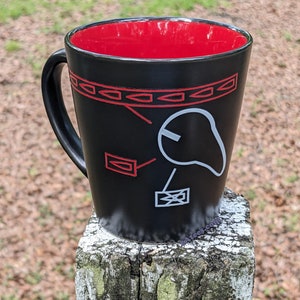 May include: Black ceramic coffee mug with a red interior and a white design of a bird with a red border. The design includes geometric shapes and lines.