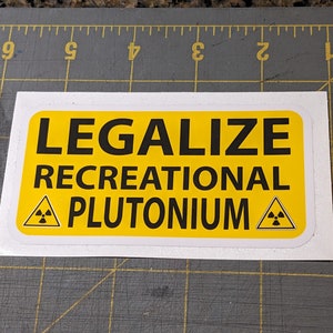 Puede incluir: Una pegatina amarilla con texto negro que dice "Legalize Recreational Plutonium" con un símbolo de radiación negro y amarillo a cada lado del texto.