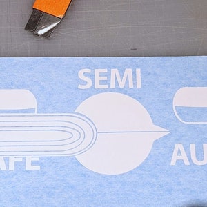 May include: A light blue sticker with white graphics and text. The text includes "SAFE", "SEMI", and "AUTO". A utility knife is in the upper left corner.