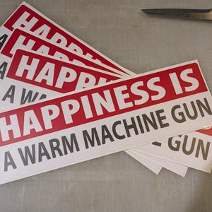 May include: A stack of white rectangular stickers with red and black text. The stickers read "HAPPINESS IS A WARM MACHINE GUN". Scissors are in the upper right corner.