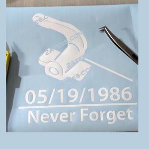 May include: A light blue vinyl sheet with a white graphic of a tape dispenser and the text "05/19/1986 Never Forget". A pair of tweezers is on the right side of the sheet. The website 2ATEES.COM is printed multiple times.