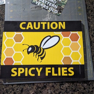 May include: A rectangular sign with a yellow honeycomb pattern background and a black border. The sign features a black and white bee illustration and the words "CAUTION SPICY FLIES" in bold yellow text.