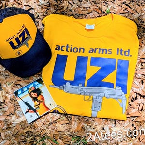 May include: A yellow t-shirt, a black and yellow trucker hat, and a small card are displayed on a bed of leaves. The shirt and hat feature the text "action arms ltd. UZI" in blue. The card shows a woman holding a gun.