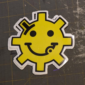 May include: A yellow smiley face sticker with a gear design around the outside. The smiley face has a black outline and a black tongue sticking out. The sticker is on a gray background.