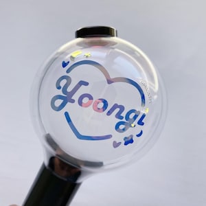 May include: A clear, spherical light stick with a black handle. The light stick features the name "Yoongi" in a heart-shaped design with blue and pink accents. Small star and heart shapes are scattered around the design.