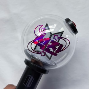 May include: A clear, spherical light stick with a black handle. The light stick features a holographic design with the word "FIRE" in purple and pink, along with stars and a crescent moon. The top has a black cap with a red button.